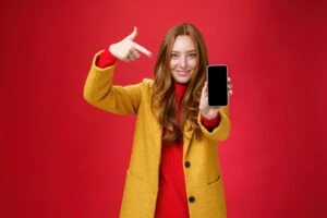 pick this phone you never regret portrait friendly looking attractive confident redhead glamour girlfriend yellow coat showing smartphone pointing mobile as smiling broadly